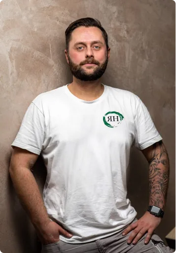 Mann mit kurzem braunem Haar, Bart und Tattoo auf dem Unterarm steht vor grauer Wand.