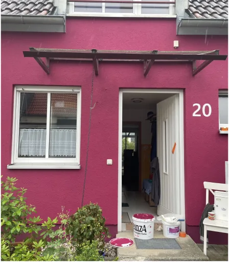 Hausfassade mit pinker Wand, weißem Fenster und geöffneter Tür, davor Farbeimer und Pflanzen.