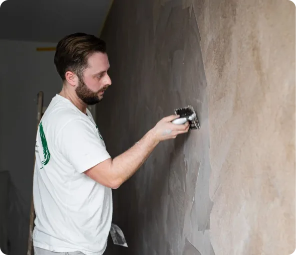 Mann in weißem T-Shirt trägt mit einem Spachtel Material an der Wand auf.