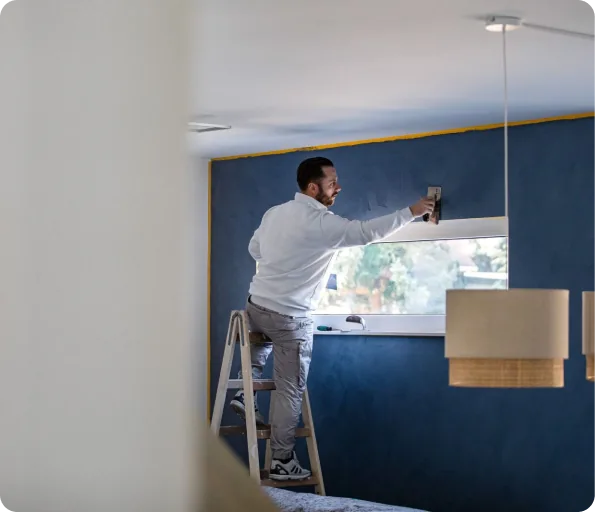 Mann auf Leiter streicht blaue Wand über Fenster in modernem Raum.
