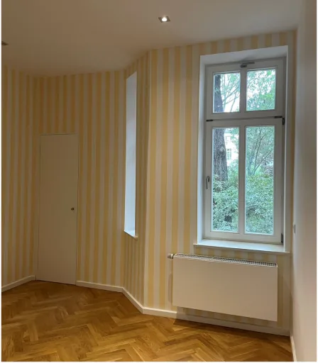 Zimmer mit gelb-weiß gestreiften Wänden, Holzparkettboden, kleinem Fenster und Heizkörper.