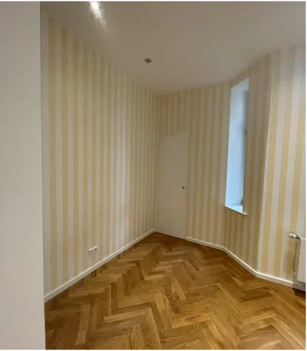 Leerer Raum mit gelb-weiß gestreiften Wänden, Holzparkettboden und kleinem Fenster.