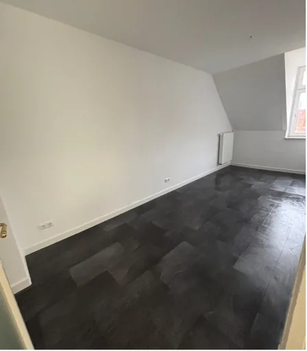 Leerer Raum mit dunklem Boden, weißen Wänden, Fenster und Heizung unter Dachschräge.