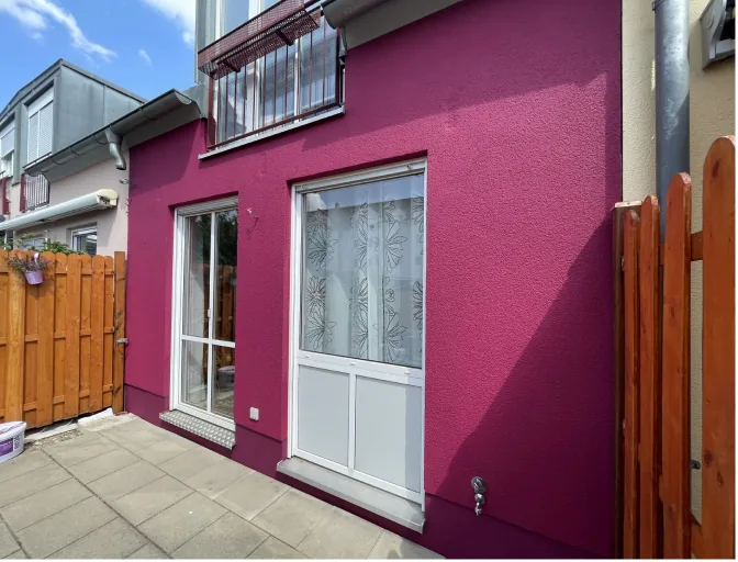 Hausfassade in kräftigem Pink mit weißer Tür, Fenster und Holzzaun bei sonnigem Himmel.